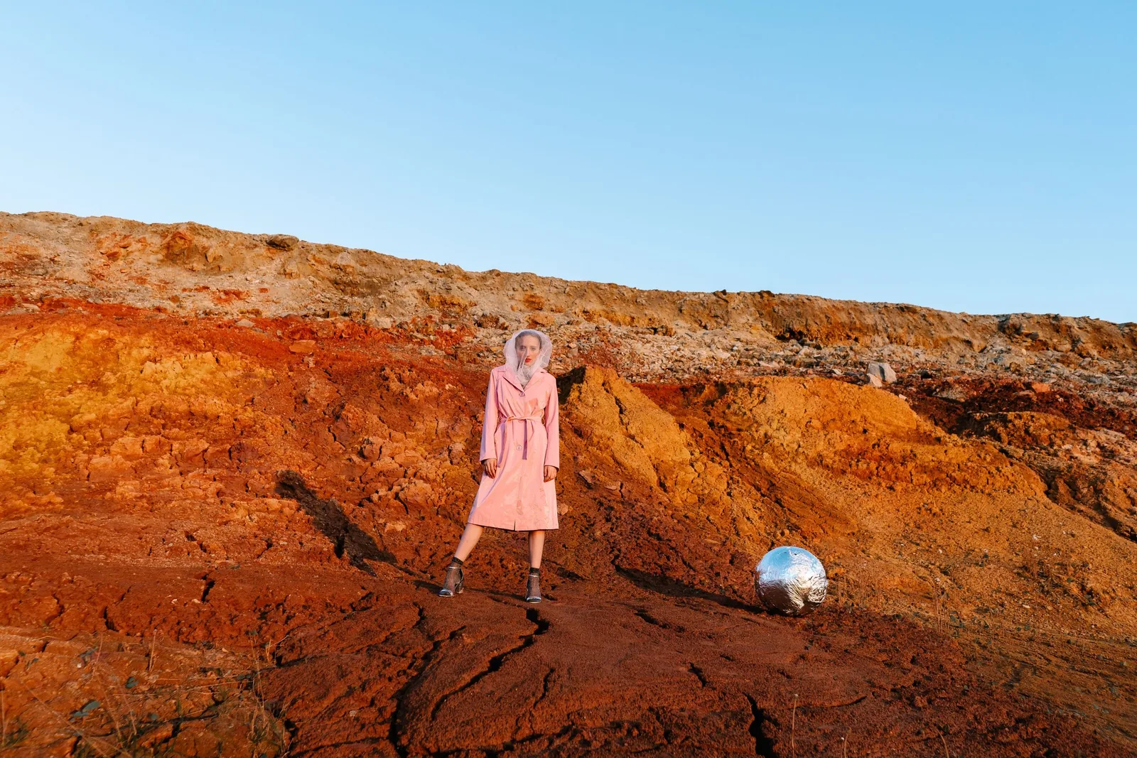 Model in a pink coat standing on red earth with a mirror ball at her feet